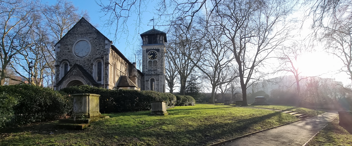 An old church in winter, viewed across a small churchyard with a few gravestones. The sun is low in the sky over a bock of flats.